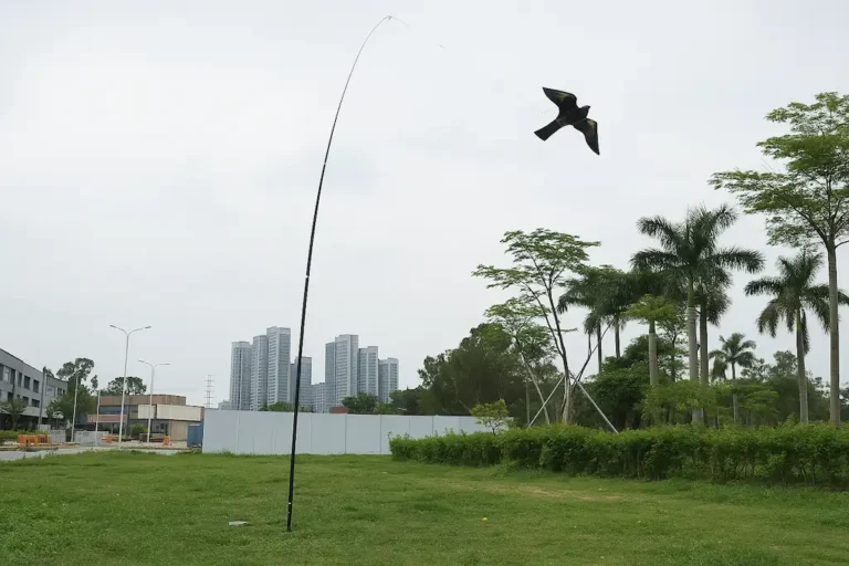 Pájaro espantapájaros atado con un hilo en una varilla vertical clavada al suelo, muy larga y flexible que le da movimiento, este ahuyentador en concreto tiene forma de ave rapaz para ahuyentar palomas, está en un descampado con hierba y de fondo palmeras y rascacielos